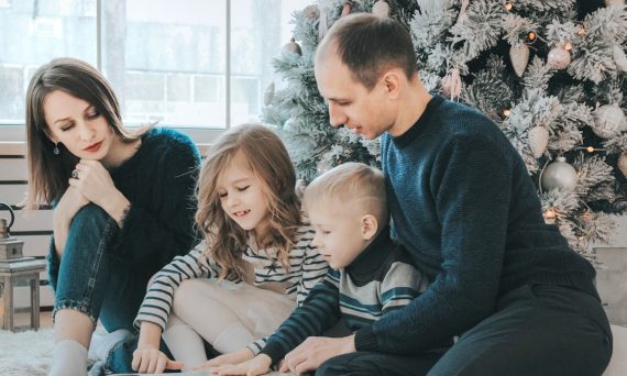 girl and boy reading book sitting between man and woman beside Christmas tree